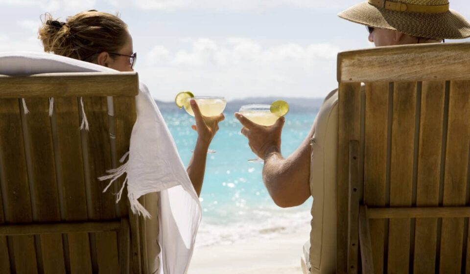 people sit in two wooden beach chairs facing the Caribbean ocean holding drinks and making a toast